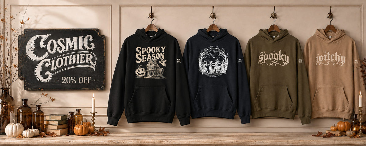 Cosmic Clothier fall collection — four witchy hoodies on brass hooks beside a vintage apothecary display with candles, amber bottles and pumpkins